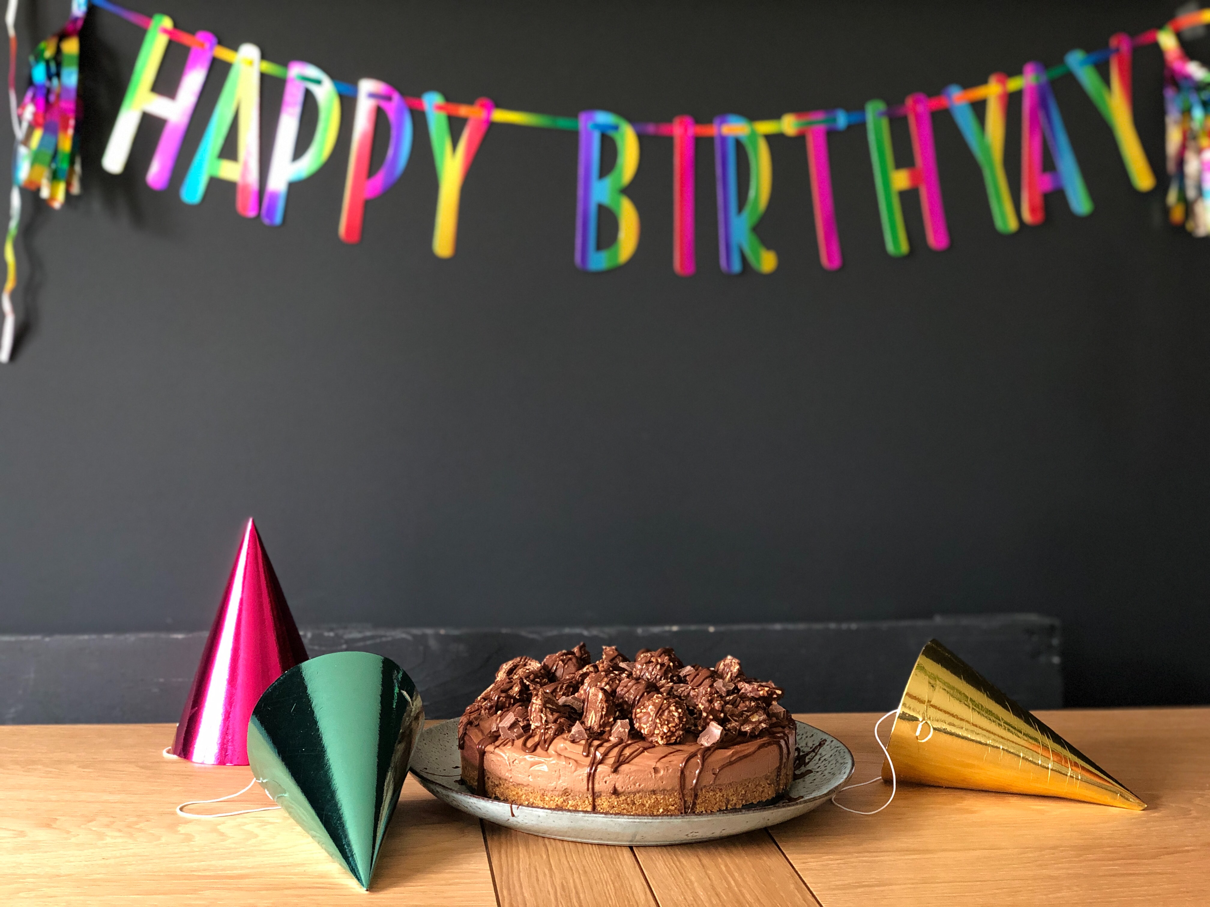 a rainbow banner reads "happy birthday," below this a chocolate cake and three party hats, two of which have fallen over